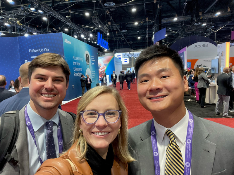 Three people at a conference with booths labeled "Alcon" and "Apellis" in the background.